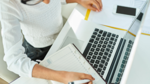 Upshot of girl in an office with laptop in front of her writing in her calendar.