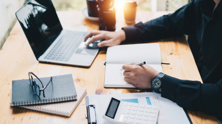 Person working at a wooden desk with a laptop, notebook, calculator, and documents
